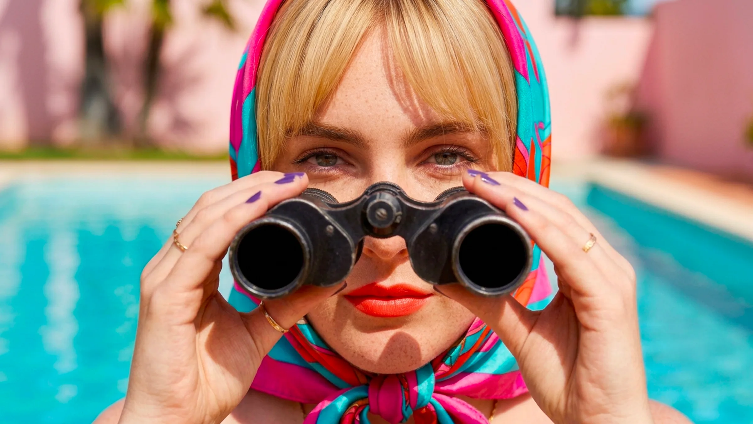 Femme au foulard coloré observant à la jumelle au bord d'une piscine, face à l'objectif